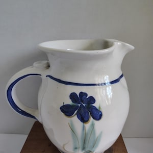 May include: White ceramic pitcher with a blue floral design. The pitcher has a rounded body, a handle, and a spout. A blue band encircles the top. The floral design features a blue flower with green leaves.