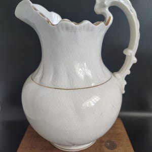 May include: A white ceramic pitcher with a ruffled rim and ornate handle. The pitcher has a bulbous body and is accented with gold trim. The pitcher is likely vintage or antique, and is displayed on a wooden surface.
