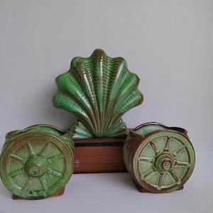 May include: A collection of ceramic planters in shades of green and brown. The set includes two wagon wheel planters and a large shell-shaped planter. The planters are likely vintage or antique, adding a touch of rustic charm to any space.
