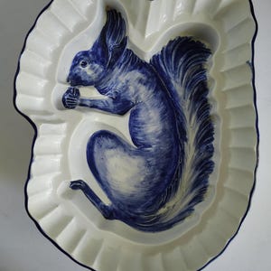 May include: A cream-colored ceramic baking mold with a scalloped edge and a blue painted design of a squirrel holding a nut. The interior features a raised, three-dimensional relief of the squirrel, with blue detailing on the tail and body.