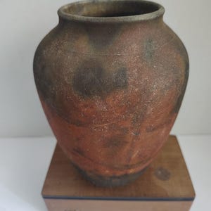 May include: A rustic, earthenware pot with a rounded body and a wide opening. The pot has a textured surface with shades of brown, red, and black. It sits on a small, wooden box. This handcrafted vessel is suitable for home decor.