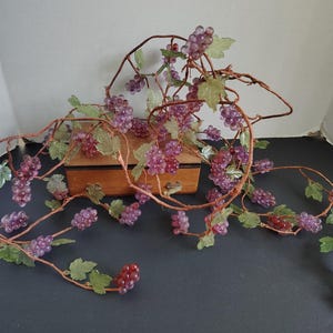May include: A decorative artificial grapevine garland featuring translucent purple plastic grapes and green fabric leaves attached to brown twisted stems, draped over a rectangular wooden box with a dark horizontal accent line.