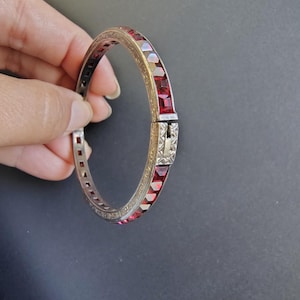 May include: A hinged bangle bracelet featuring a metallic band with an etched pattern and a series of square-cut, deep red stones set along the outer edge. The bracelet includes a secure metal clasp mechanism that connects the two ends together.