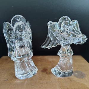 May include: Two clear glass angel figurines. One angel has the word "Hope" etched on its front. The other angel has the word "Generosity" etched on a banner. Both angels have wings and are standing on a wooden surface.