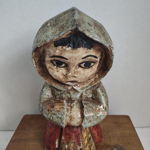 May include: A hand-carved wooden figurine of a person wearing a hooded cloak. The figurine has a serene expression, with dark eyes and a closed mouth. The cloak is painted in shades of green and brown, and the skirt is red and yellow. The figurine is placed on a wooden surface.