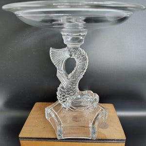 May include: A clear glass serving dish with a fish-shaped support. The dish has a wide, shallow bowl and sits atop a detailed fish sculpture. The base is a clear, geometric shape, and the dish is presented on a wooden box.