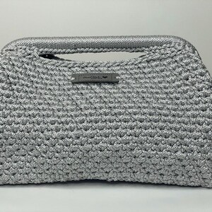 May include: A silver crocheted handbag with a handle and a small rectangular tag. The bag has a textured, woven appearance and a rounded shape. The tag reads "Hand Made".