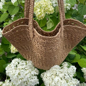 May include: A woven brown tote bag with two handles. The bag has a wide, open design and is made of a textured material. The background features white hydrangea flowers and green leaves.