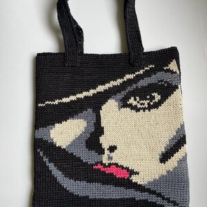 May include: Black crochet tote bag featuring a stylized face design. The face is rendered in shades of black, gray, and cream, with a pop of pink for the lips. The bag has two black handles and a rectangular shape.