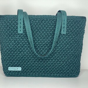 May include: Teal-colored tote bag with a textured, woven design. The bag features matching teal leather-like handles and a small label that reads "Hand Made". The bag is a medium size and appears sturdy.