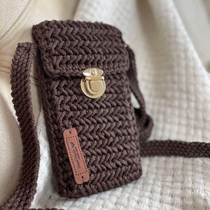 May include: A dark brown, handmade crochet crossbody phone bag with a gold-colored clasp. The bag features a textured stitch pattern and a matching strap. A small leather tag with the words "Hand Made" is attached.