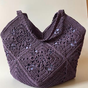 May include: A handcrafted, purple crochet tote bag. The bag features a geometric floral pattern and two handles. The bag's open weave design is visible, and the bag is made of a soft, durable material.