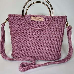 May include: A mauve-colored, handmade crochet handbag with gold-colored metal handles and a detachable shoulder strap. The bag features a rectangular shape and a textured stitch pattern. A small tag reads "Hand Made with Love."