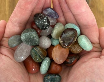 May include: A handful of polished gemstones in various colors, including red, green, brown, and purple. The stones are smooth and rounded, with a natural, earthy appearance. The gemstones are held in open hands.
