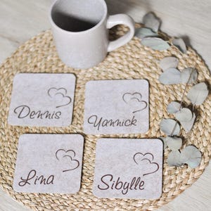 Può includere: Una tazza in ceramica beige è appoggiata su un sottobicchiere intrecciato, con quattro sottobicchieri quadrati in feltro grigio. Ogni sottobicchiere ha un nome in scrittura marrone: Dennis, Yannick, Lina e Sibylle, insieme a un disegno di nuvola.