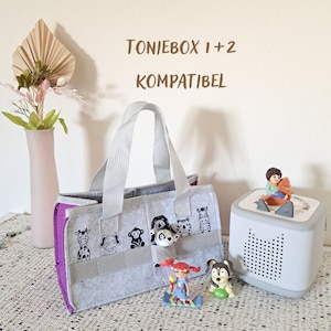 Peut inclure: Un sac de rangement en feutrine gris et violet avec des illustrations d'animaux, doté de deux anses gris clair et d'une petite poche avant. Le texte TONIEBOX 1+2 KOMPATIBEL est visible au-dessus. Une enceinte gris clair et deux figurines.