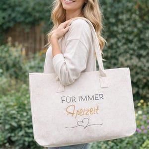 Könnte beinhalten: Beige Tragetasche mit dem deutschen Text "FÜR IMMER Freizeit" in schwarzer und goldener Schrift, mit einem Herzdesign. Die Tasche hat zwei Griffe und wird über der Schulter einer Person getragen.
