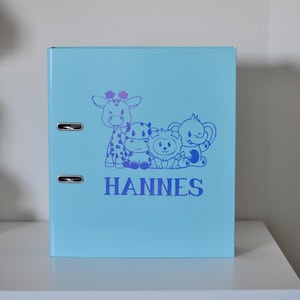 Puede incluir: Carpeta azul claro con una ilustración morada de cuatro animales de dibujos animados: una jirafa, un hipopótamo, un león y un elefante. El nombre "HANNES" está impreso en morado. La carpeta tiene dos anillas metálicas.