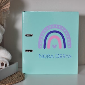 Puede incluir: Carpeta de color turquesa claro con un diseño de arcoíris en morado, rosa y azul. El nombre "NORA DERYA" está impreso en azul debajo. La carpeta tiene dos anillas metálicas.