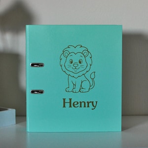 Puede incluir: Un archivador turquesa con una ilustración de león dorado y el nombre "Henry" impreso debajo. El archivador tiene dos anillas metálicas y está diseñado para organizar documentos. El diseño es sencillo y apto para niños.
