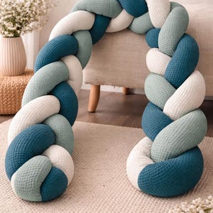 Könnte beinhalten: Ein geflochtener Baby-Bumper in Blau-, Türkis- und Weißtönen. Der Bumper besteht aus einem strukturierten Stoff und soll das Baby vor den harten Oberflächen des Kinderbetts schützen. Der Bumper liegt auf einem neutralen Teppich.
