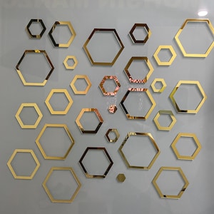Puede incluir: Una colección de decoración mural geométrica hexagonal dorada de varios tamaños. Los hexágonos están dispuestos en un patrón disperso sobre un fondo gris claro, creando un diseño decorativo moderno.