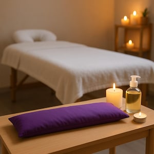 Puede incluir: Una almohada rectangular de aromaterapia morada descansa sobre una mesa de madera, junto a una vela encendida, una pequeña vela de té y una botella de aceite. Una camilla de masaje con una sábana blanca está en el fondo.