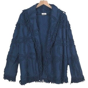 May include: A navy blue open-front jacket with a textured, patterned design and fringe trim. The jacket is hanging on a wooden hanger. The fabric has a woven appearance with a decorative pattern.