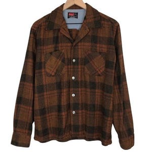May include: A brown and black checked button-up shirt with two chest pockets. The shirt features a classic collar and is made of a thick, warm material. The shirt is buttoned up and hanging on a wooden hanger.
