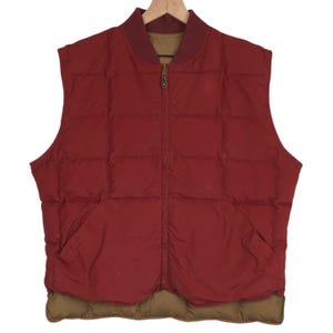 May include: A reversible, sleeveless puffer vest in a deep red color with a brown collar and hem. The vest has a full-length zipper and two front pockets. The vest is hanging on a wooden hanger.