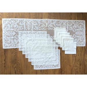 Puede incluir: Un camino de mesa de encaje blanco y manteles individuales y servilletas a juego están dispuestos sobre una superficie de madera. El camino de mesa y los manteles individuales presentan un diseño floral y de volutas detallado. Las servilletas son blancas lisas.