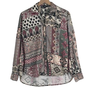 Puede incluir: Una camisa de manga larga con botones que presenta un patrón de retazos multicolor en tonos rojo apagado, verde oliva, crema y negro. La prenda tiene un cuello estándar, una tapeta de botones frontal y un dobladillo curvo con costuras visibles.