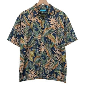 Peut inclure: Une chemise à manches courtes avec un motif de feuilles tropicales. La chemise présente un fond bleu marine avec des feuilles vertes, beiges et marron. Elle est suspendue à un cintre en bois. Style boutonné.
