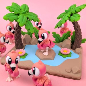 May include: A miniature diorama with pink flamingo figures, each with large white eyes and black beaks. The scene includes green palm trees, a blue pond, and pink flowers. The background is a solid pink colour.