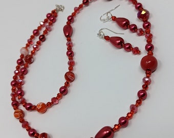 May include: A red beaded necklace and matching earrings. The necklace is made with red beads of various sizes and shapes, including round, oval, and teardrop. The earrings are teardrop shaped and dangle from silver hooks.