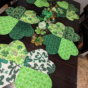 May include: Five green shamrock-shaped placemats with different patterns of shamrocks and swirls. The placemats are on a dark brown wooden table.