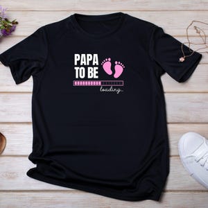 Peut inclure: T-shirt noir avec l'inscription blanche "PAPA TO BE", des empreintes de pieds roses et une barre de chargement. Le mot "loading..." est écrit en dessous dans une police script. Un cadeau parfait pour un nouveau papa.