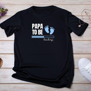 Peut inclure: T-shirt noir avec l'inscription blanche "PAPA TO BE", des empreintes de pieds de bébé bleues et une barre de chargement. Le mot "loading..." est en dessous. Un t-shirt décontracté pour les futurs parents.