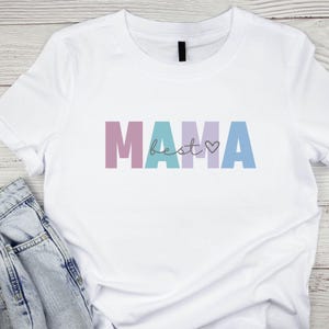 Könnte beinhalten: Weißes T-Shirt mit dem Wort "MAMA" in lilafarbenen, türkisfarbenen und blauen Blockbuchstaben. Das Wort "best" ist in einer Schreibschrift zwischen den Buchstaben geschrieben, mit einem kleinen Herzsymbol. Eine blaue Jeans liegt neben dem T-Shirt.
