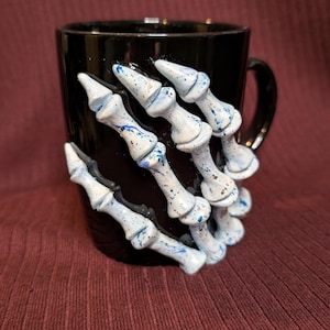 May include: Black ceramic mug with a white and blue speckled skeleton hand design. The hand is sculpted to appear as if it is gripping the mug. The mug has a black handle and is set against a burgundy background.