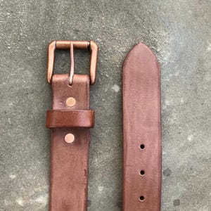 May include: A brown leather belt with a copper-colored buckle and rivets. The belt has a classic design with a pointed end and multiple holes for adjustment. The buckle is rectangular and securely attached to the belt.