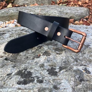 May include: A black leather belt with a copper-colored buckle and rivets. The belt is lying on a gray stone surface, showcasing its simple design and quality craftsmanship. The buckle is square and the rivets are circular.