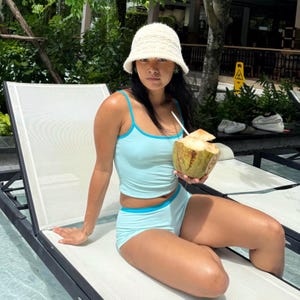 May include: A light blue two-piece swimsuit with turquoise trim. The outfit is paired with a white crocheted bucket hat. A coconut with a straw is held in the hand.