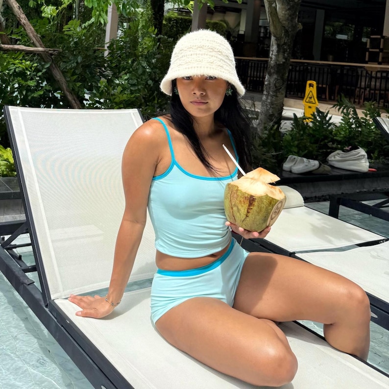 May include: A light blue two-piece swimsuit with turquoise trim. The outfit is paired with a white crocheted bucket hat. A coconut with a straw is held in the hand.