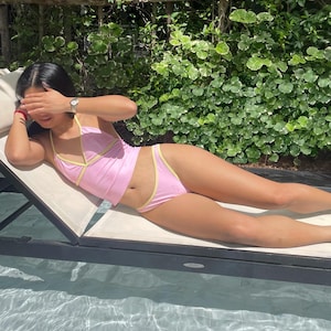 May include: Pink bikini set with yellow trim. The top is a halter style, and the bottoms are high-waisted. The swimwear is worn while relaxing on a lounge chair near a pool.