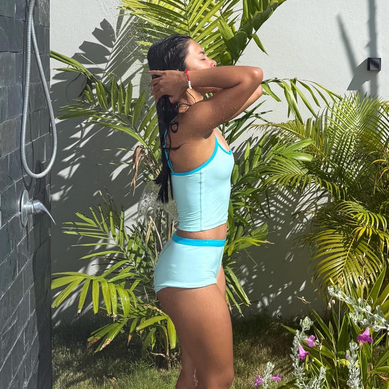 May include: A light blue two-piece swimsuit with a contrasting blue trim. The top is a crop top style with thin straps, and the bottoms are high-waisted shorts. The swimsuit is worn outdoors near a shower head and tropical plants.