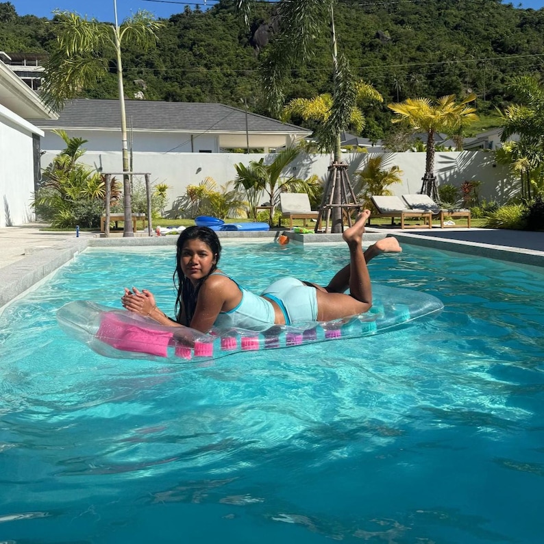 May include: A woman wearing a light blue swimsuit and lying on a clear inflatable pool float with a pink headrest and purple and pink supports in a swimming pool. The pool is surrounded by a patio, trees, and a house.