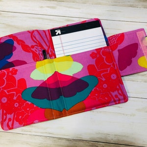 May include: A pink fabric notebook cover with a colorful butterfly design. The cover has a pocket on the outside and a pen holder on the inside. The notebook cover is open to reveal a lined notepad.