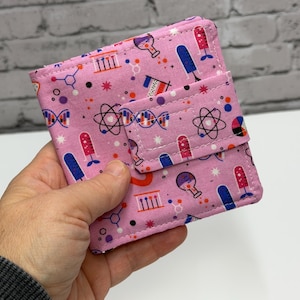 May include: Pink fabric wallet with a science-themed pattern. The pattern includes images of atoms, DNA strands, and laboratory equipment in blue, red, and white. The wallet has a flap closure.