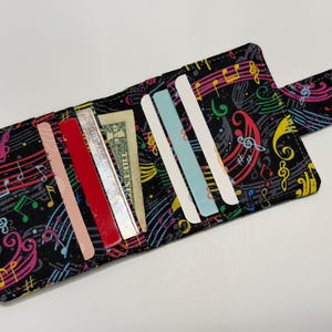 May include: Open fabric wallet with a black background and a colorful musical note pattern. The wallet is open, revealing several credit cards and a dollar bill. A Velcro closure is visible.